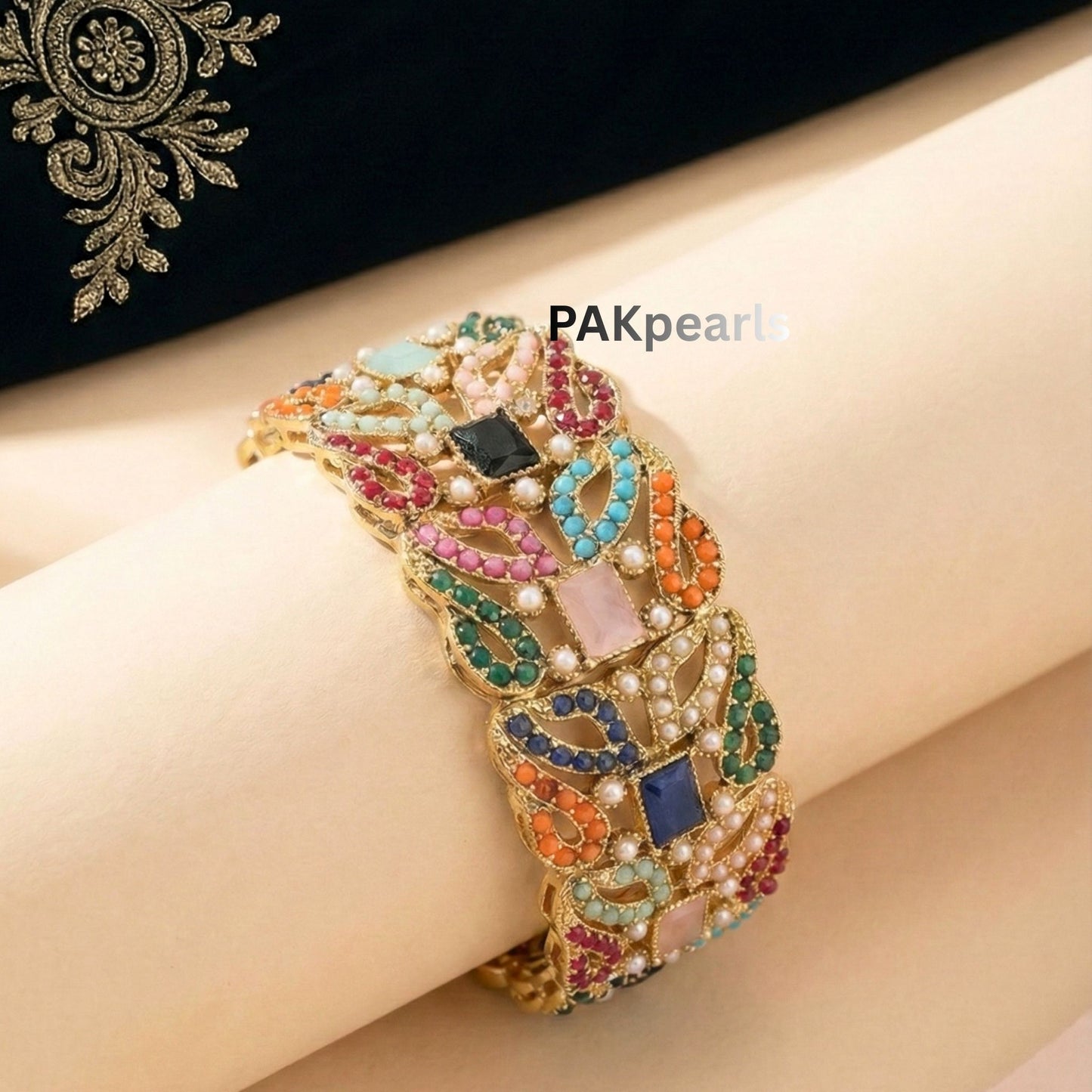 Pair of wide Nauratan gold-plated bangles featuring multi-colored stones and pearl accents displayed on a wooden stand.