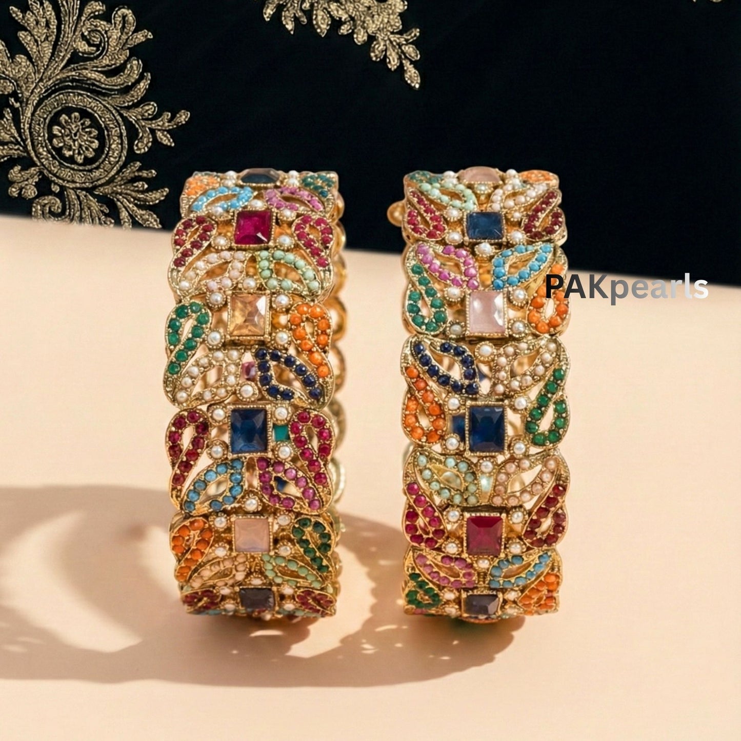 Pair of wide Nauratan gold-plated bangles featuring multi-colored stones and pearl accents displayed on a wooden stand.