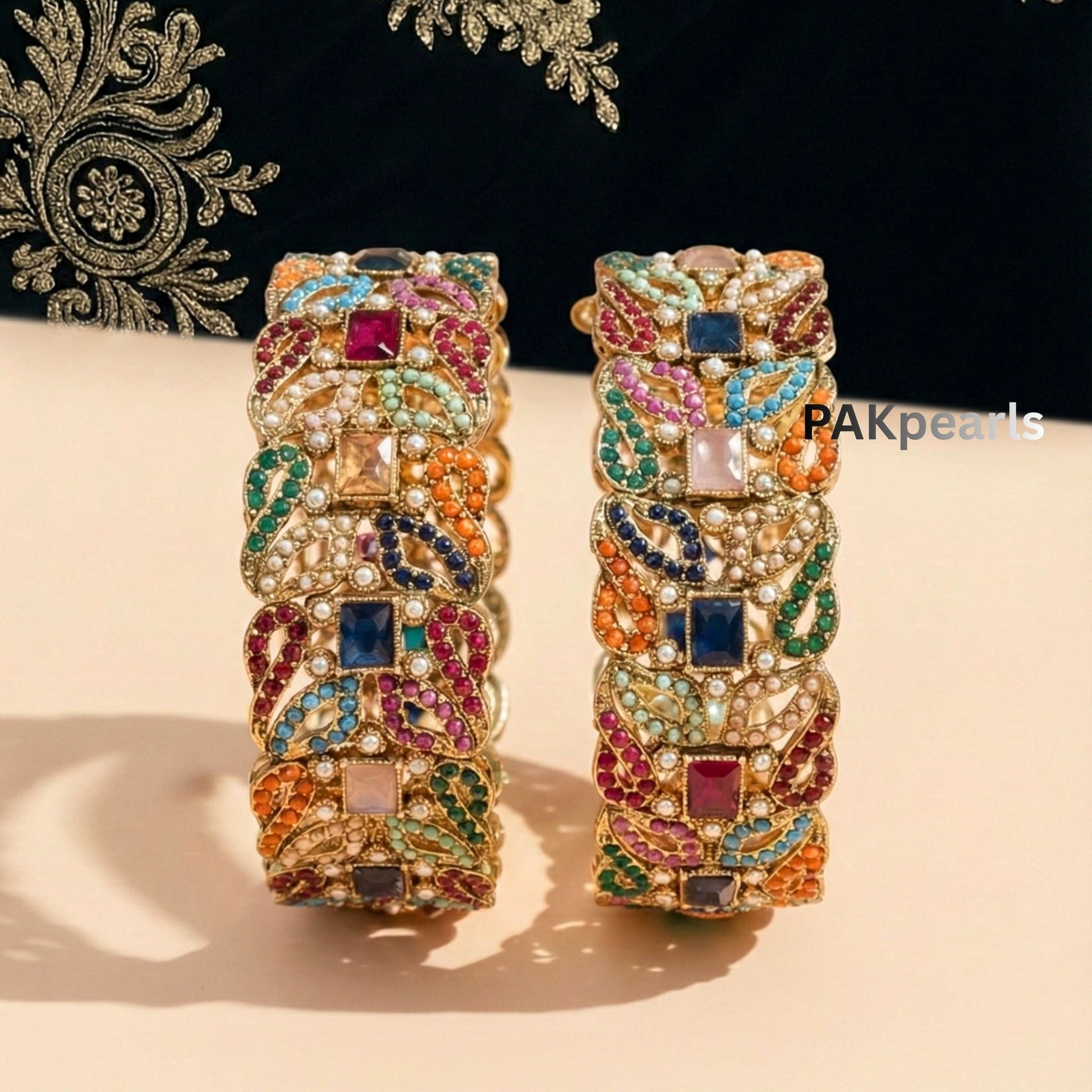 Pair of wide Nauratan gold-plated bangles featuring multi-colored stones and pearl accents displayed on a wooden stand.