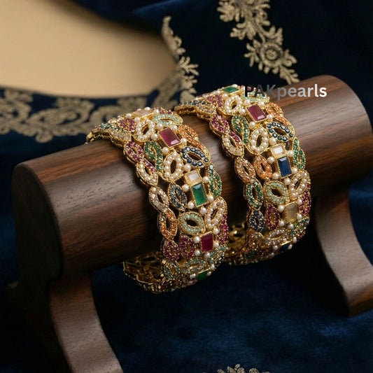 Pair of wide Nauratan gold-plated bangles featuring multi-colored stones and pearl accents displayed on a wooden stand.